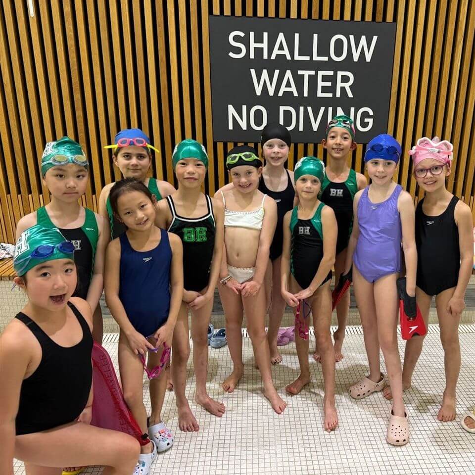 A group of twelve children in swimsuits and swim caps stand smiling by a pool. A sign behind them reads "SHALLOW WATER NO DIVING." Vertical wooden slats cover the wall in the background.