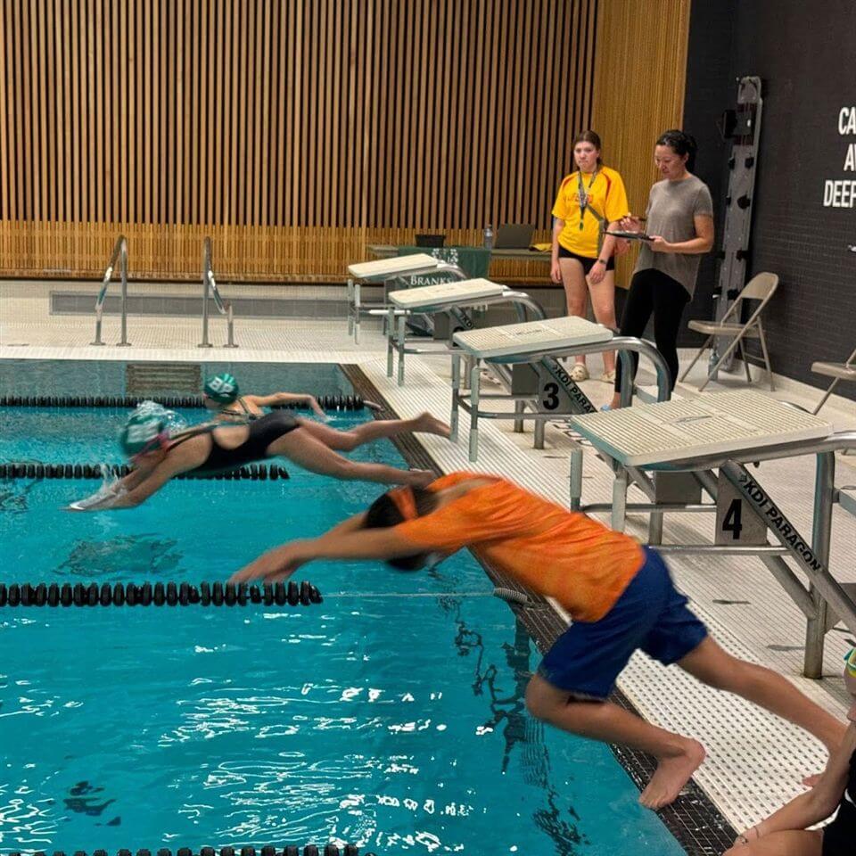 Three swimmers dive into a pool from starting blocks while two women, one in a yellow shirt, stand by the poolside observing. The swimming pool has lane dividers and a wooden wall in the background.