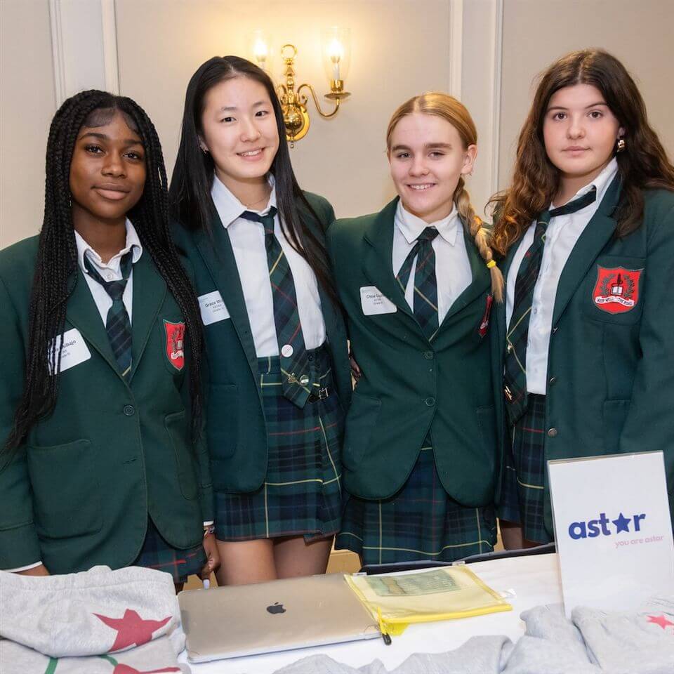 Four high school girls in green blazers and plaid skirts stand together, smiling at a table with sweatshirts and a sign that reads "ast*r you are online" in front of them.
