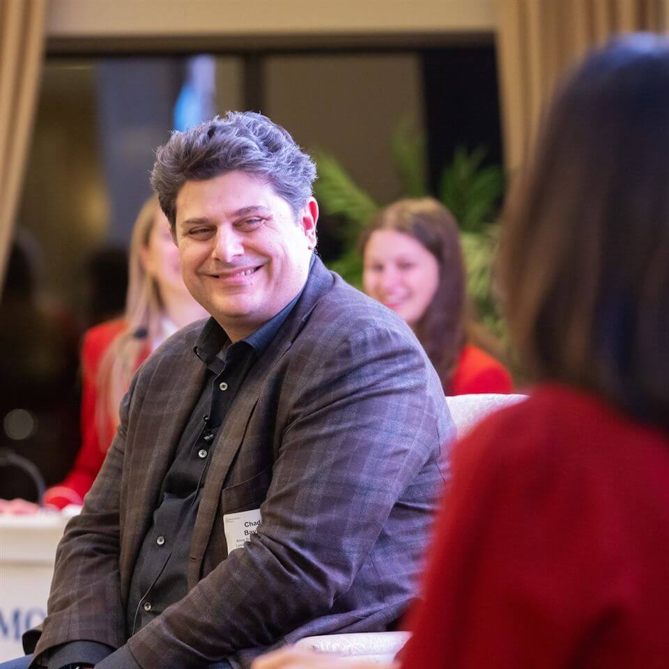 A man in a dark plaid jacket sits and smiles at a woman in the foreground, during an indoor event with other people blurred in the background. The mood appears friendly and relaxed.