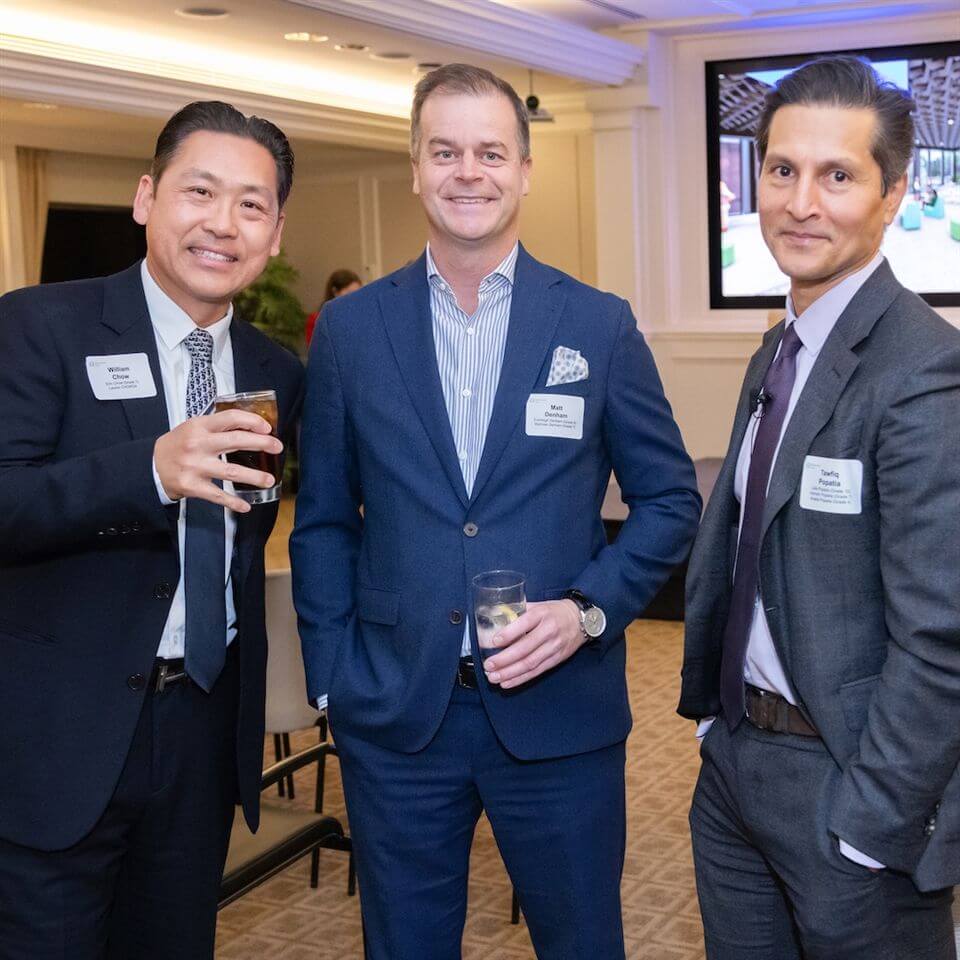 Three men in business suits stand together at an indoor event, smiling at the camera. Two hold drinks, and all wear name tags. A presentation screen and chairs are visible in the background.
