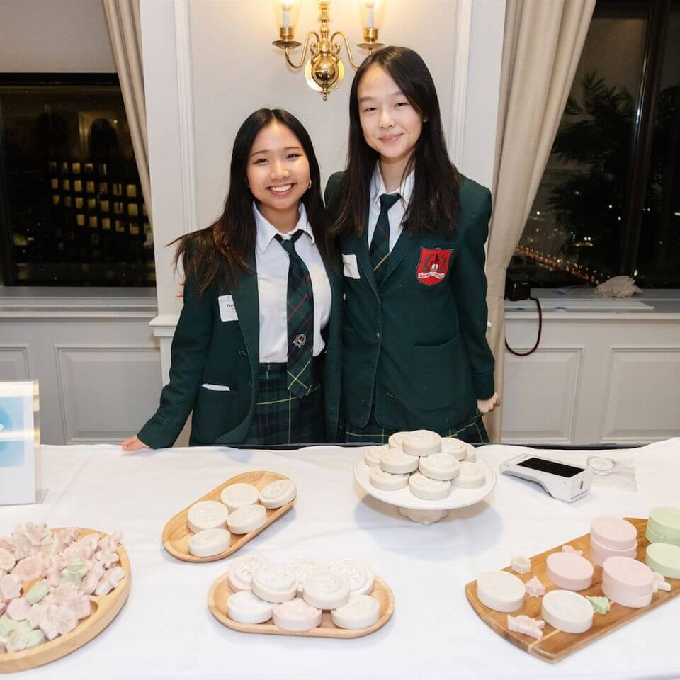 Two girls in green school uniforms stand behind a table displaying neatly arranged handmade soaps on wooden trays. They are smiling in a well-lit room with elegant decor and large windows in the background.