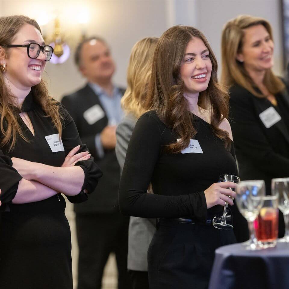 A group of professionally dressed people stand indoors, smiling and talking at a social event. A woman in the foreground holds a glass, with others nearby, some with folded arms. Networking name tags are visible on their outfits.