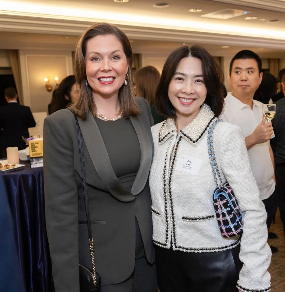 Two women posing and smiling at an indoor event, with people and a table covered with a dark cloth in the background. One woman wears a dark suit, the other a white jacket, both with shoulder-length hair.