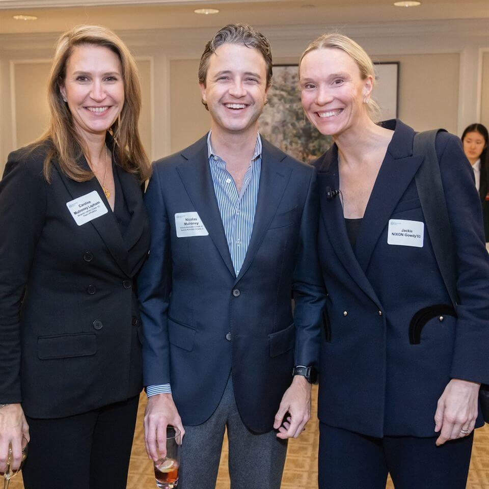 Three people dressed in business attire stand together smiling at an indoor event, each wearing name badges. One person holds a drink. There are blurred attendees and artwork visible in the background.