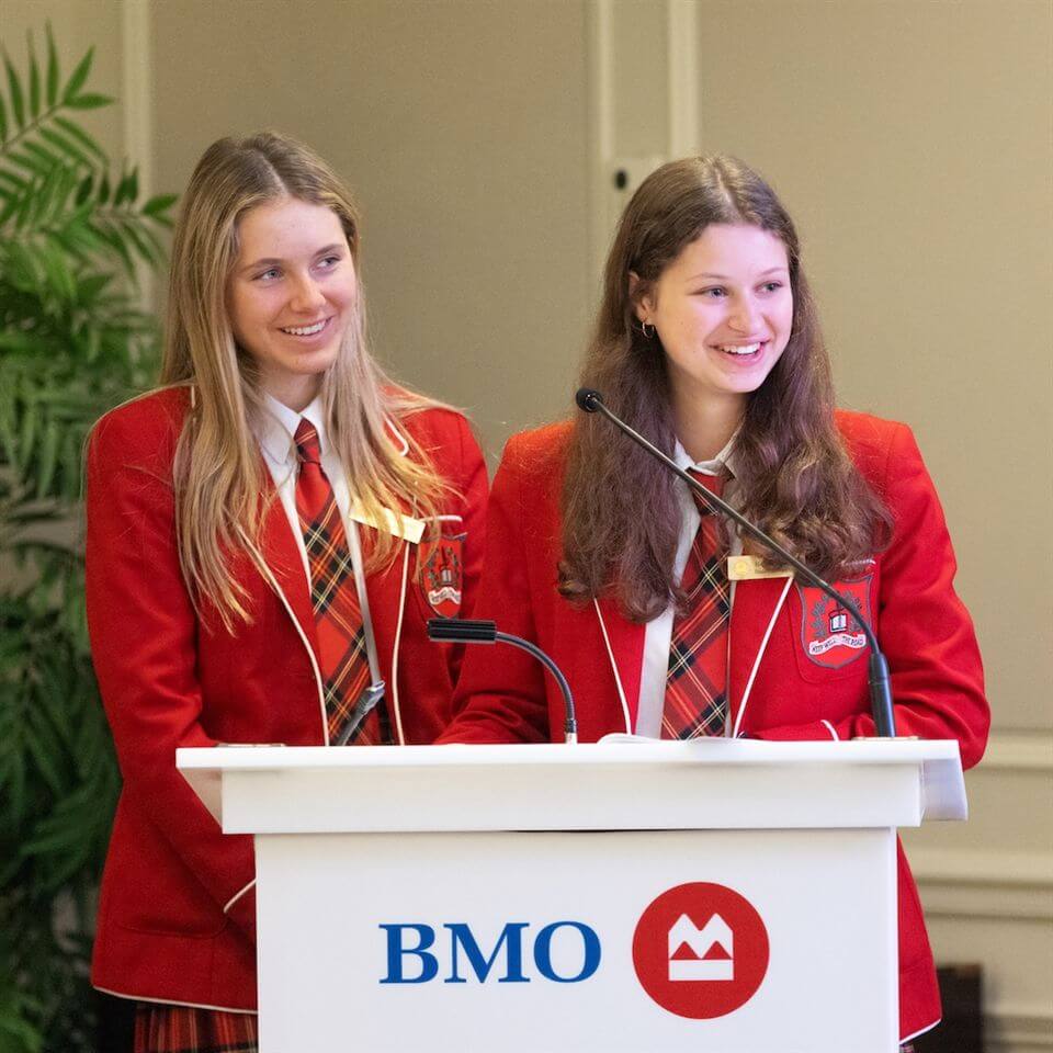 Two teenage girls in red blazers and plaid ties stand at a podium with the BMO logo, smiling while speaking or presenting in a formal indoor setting.