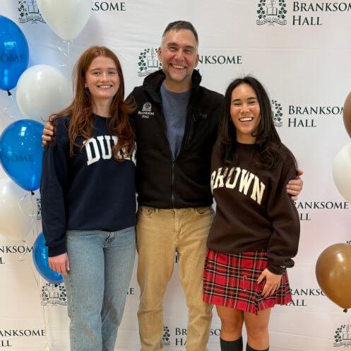 Three people stand smiling in front of a Branksome Hall backdrop with balloons. The two women wear "DUKE" and "BROWN" sweatshirts, and the man in the middle wears a black jacket and tan pants.