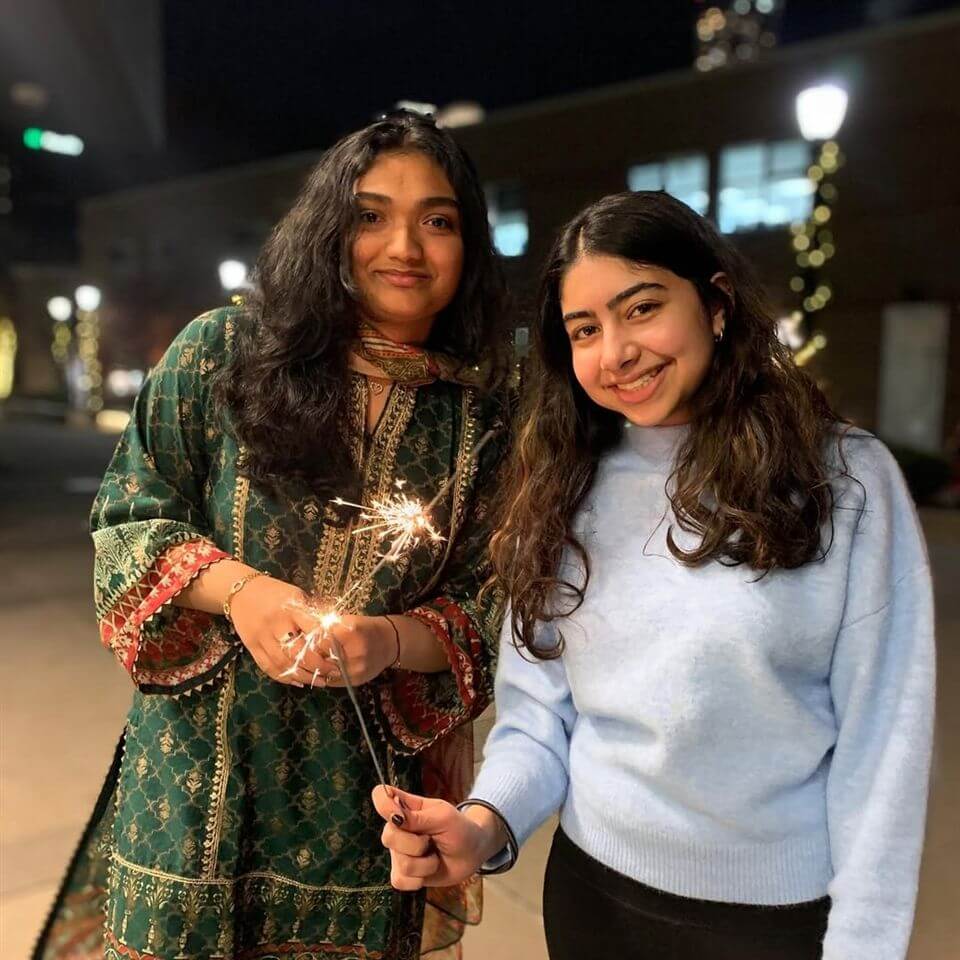 Two young women smile outdoors at night, holding sparklers. One wears a green patterned traditional outfit, and the other wears a light blue sweater. Decorative lights and buildings are blurred in the background.