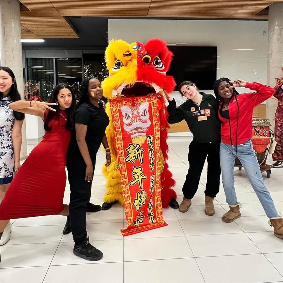 Five people pose smiling next to a colorful Chinese lion dance costume holding a bright red and gold banner with Chinese characters and “Happy New Year.” The group appears to be celebrating Lunar New Year indoors.