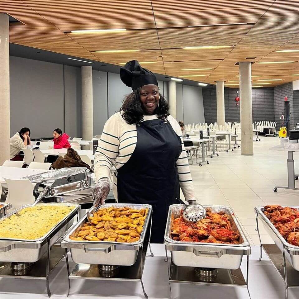 A smiling chef in a black apron and hat stands behind a buffet table with trays of food, including rice, fried plantains, and chicken, in a spacious, modern dining area.