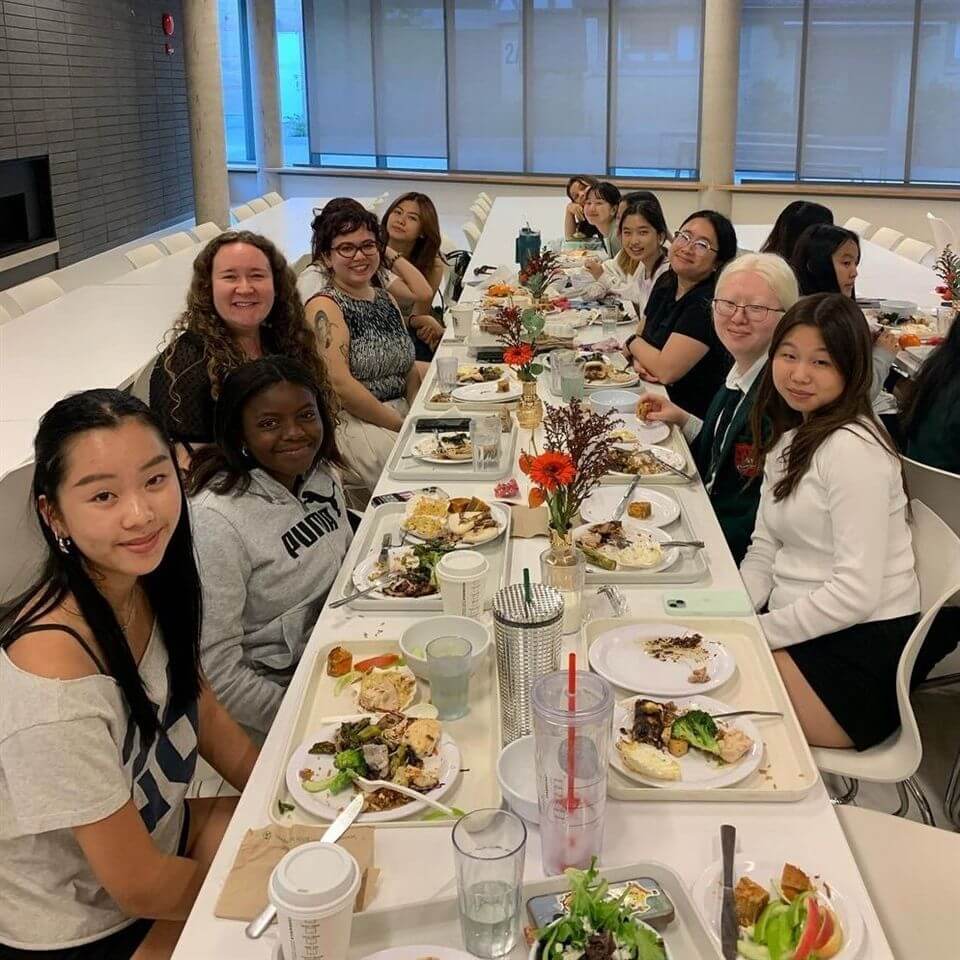 A group of women sit around a long table enjoying a meal together in a bright room. Plates of food, drinks, and small flower arrangements decorate the table. Most people are smiling and facing the camera.