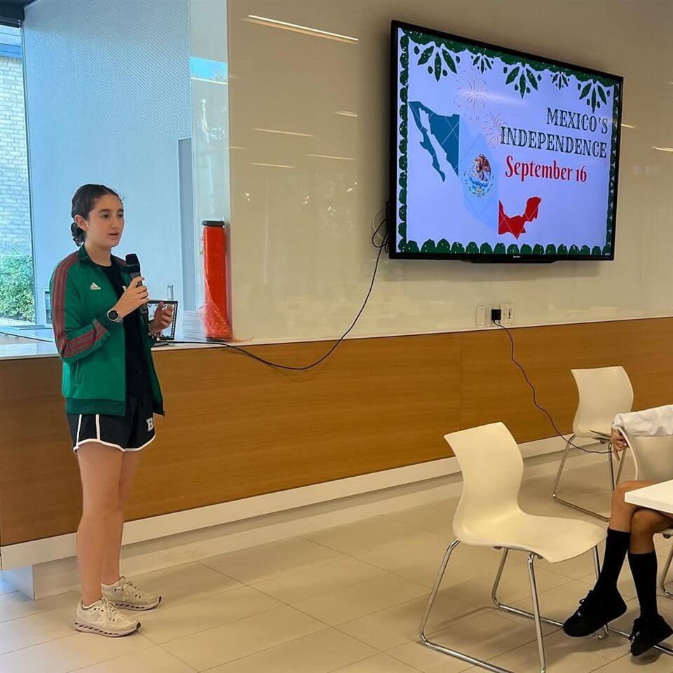 A young woman wearing a green jacket speaks into a microphone in a modern room. A screen behind her displays "Mexico's Independence, September 16" with festive graphics. White chairs and tables are visible.