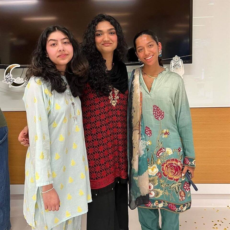 Three young women stand side by side, smiling at the camera. They are wearing colorful traditional South Asian attire, including light blue, red, and teal outfits with intricate patterns. A TV is mounted on the wall behind them.