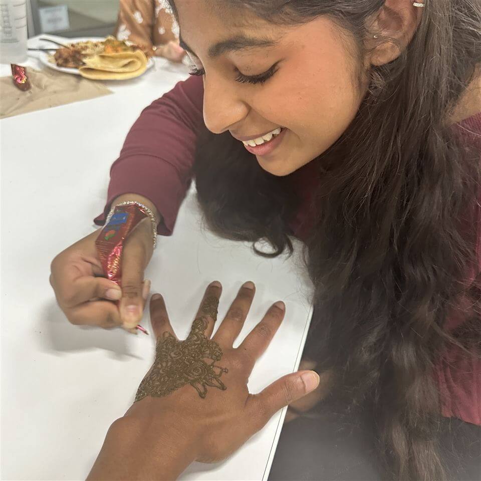 A smiling young woman with long dark hair applies an intricate henna design to another person's hand at a table. Food and drink are visible in the background.