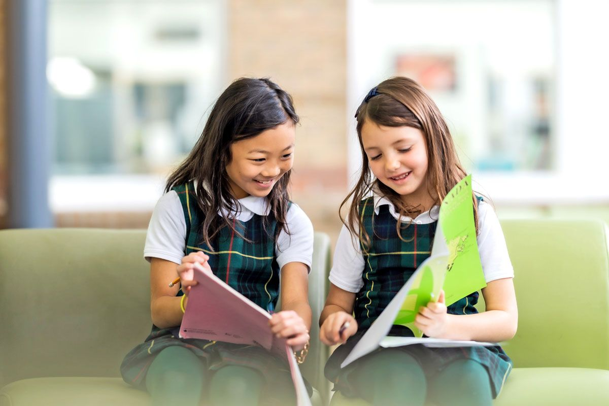 Two young girls in school uniforms sit on a green couch, smiling as they look at open folders—perhaps reviewing their IB Diploma Programme notes. The softly blurred background hints at the vibrant atmosphere of an urban campus in Toronto.