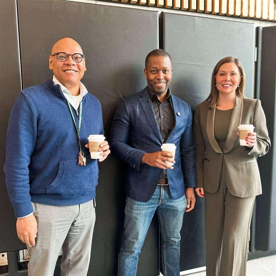 Three adults stand indoors, smiling and holding coffee cups. The man on the left wears glasses and a blue sweater, the man in the middle wears a dark blazer, and the woman on the right wears a green suit. Black panels are in the background.