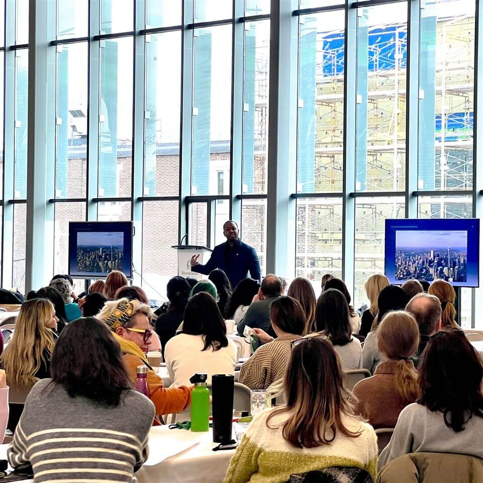 A speaker stands at a podium addressing a seated audience in a bright, modern room with large windows. Two screens display cityscape images beside the speaker. The audience listens attentively.