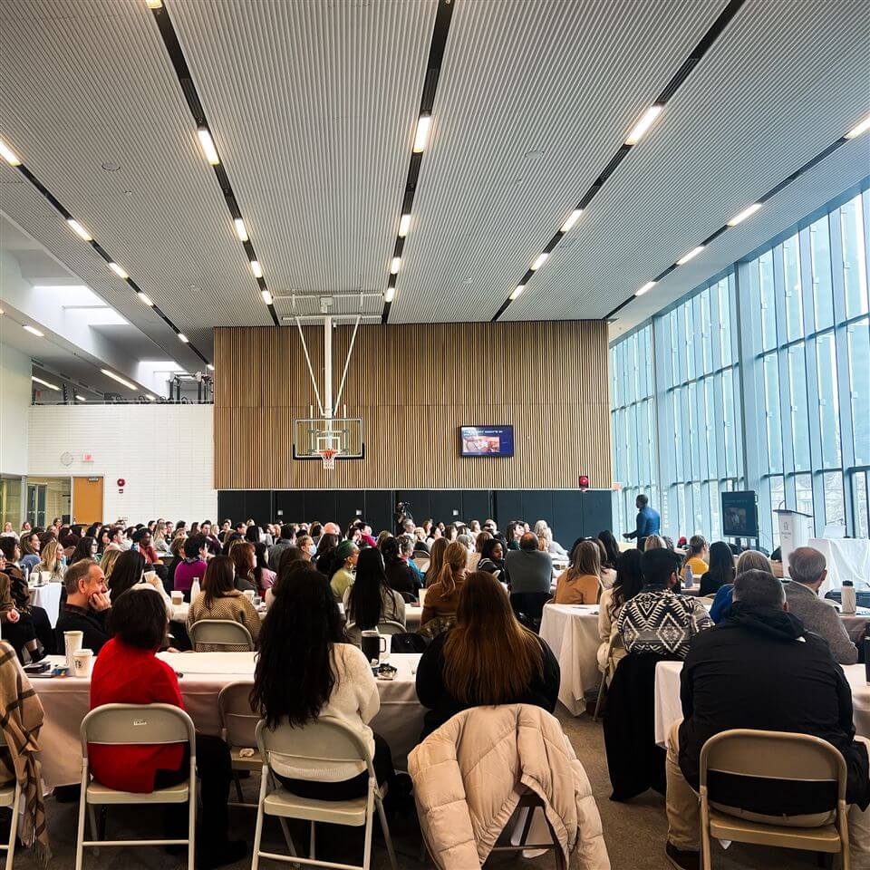 A large group of people sit at round tables in a bright, modern room with high ceilings and large windows, listening to a speaker at the front near a podium and a basketball hoop.