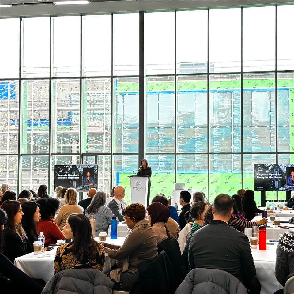 A woman speaks at a podium in front of large windows to a seated audience at round tables during a conference or seminar. Two screens display her image on either side of the stage. Construction is visible outside.