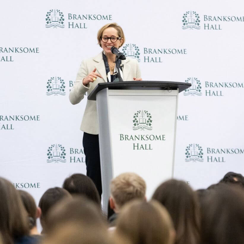 A woman in glasses speaks at a podium with a microphone in front of an audience. The podium and backdrop display the Branksome Hall logo and name.