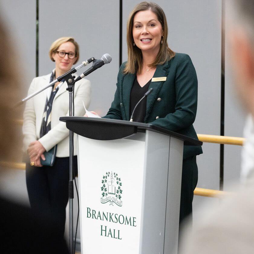 A woman in a green suit speaks at a podium labeled "Branksome Hall" while another woman stands and listens in the background. Both women are indoors at a formal event.