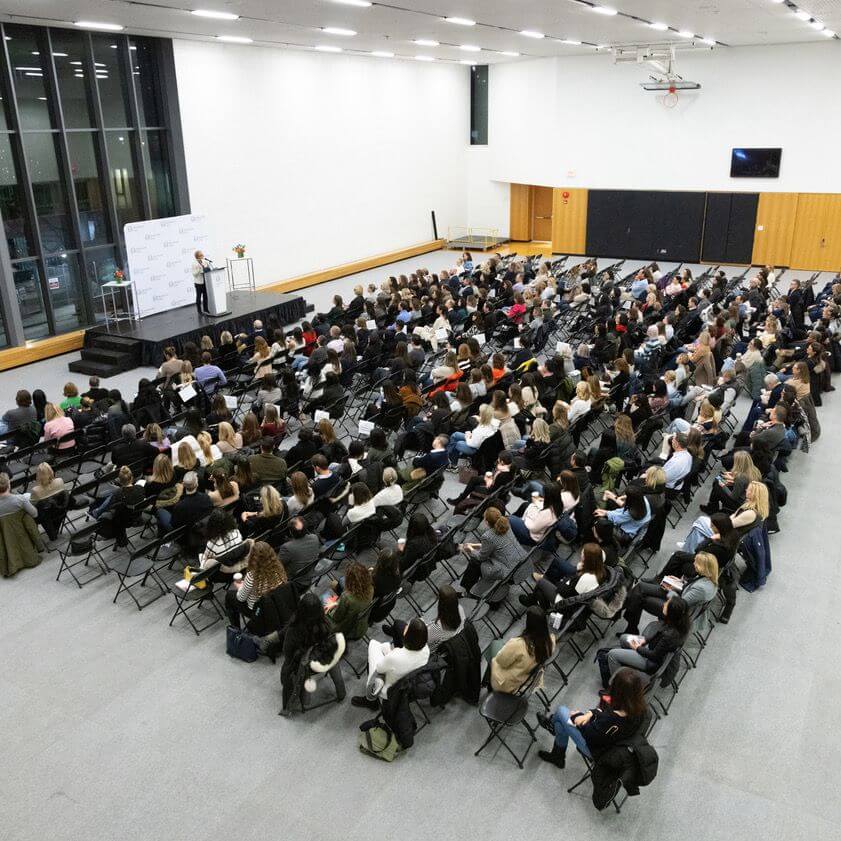 A large audience sits in rows of chairs facing a stage where a speaker stands at a podium in a bright, modern hall with high ceilings and large windows.