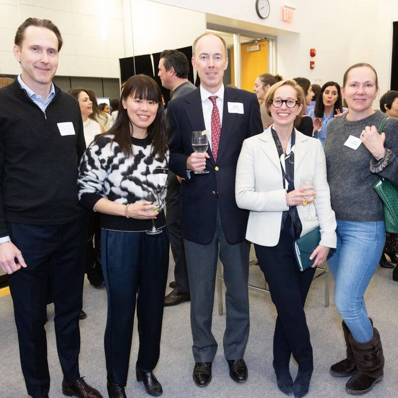 Five adults pose together at a social event, holding drinks and smiling. They wear business-casual attire and name tags, standing indoors with other attendees visible in the background.