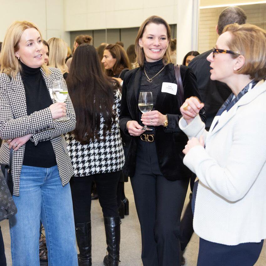 Three women stand together at a social event, holding glasses of white wine and conversing. Other people are mingling in the background, and everyone is dressed in business casual attire.