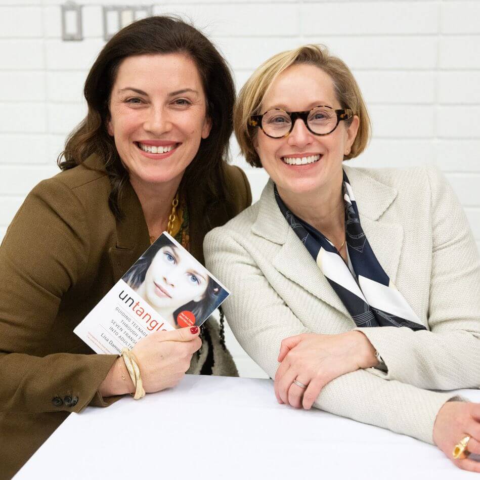 Two smiling women sit at a table. One holds up a book titled "Untangled" with a young woman's face on the cover. Both women wear blazers and glasses, and the background features a white brick wall.