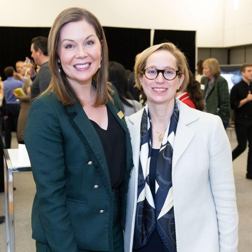 Two women standing side by side, smiling at the camera at an indoor event. One wears a dark green suit, the other a white jacket with a patterned scarf. People are visible conversing in the background.