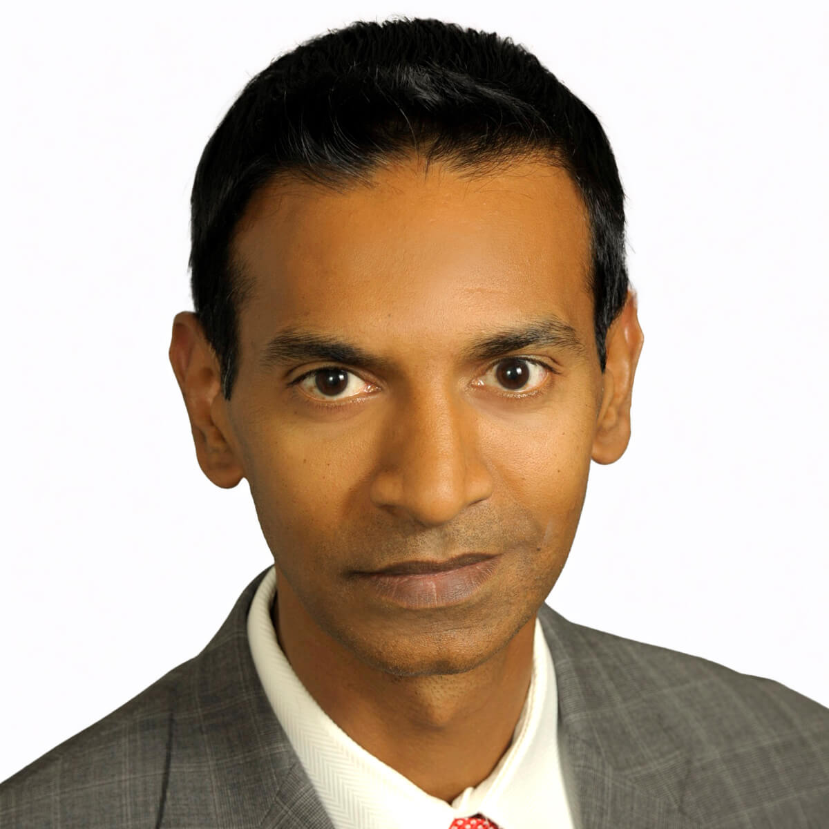 A man wearing a gray suit, white shirt, and red tie is looking directly at the camera with a neutral expression against a plain light background.