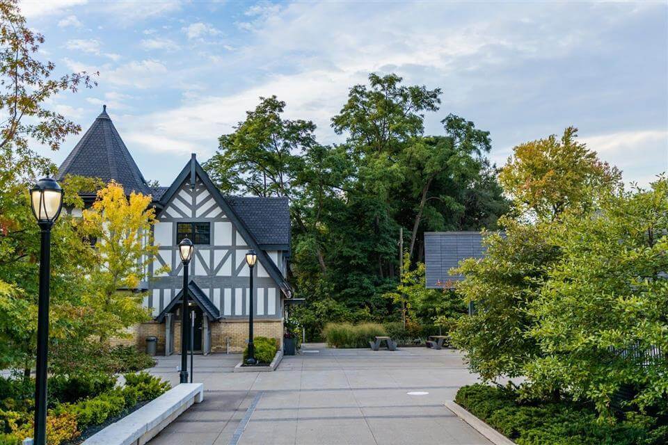A Tudor-style building with white and dark wood exterior sits beside a paved courtyard, surrounded by trees with green and yellow leaves under a partly cloudy sky—a serene setting for global education for girls.