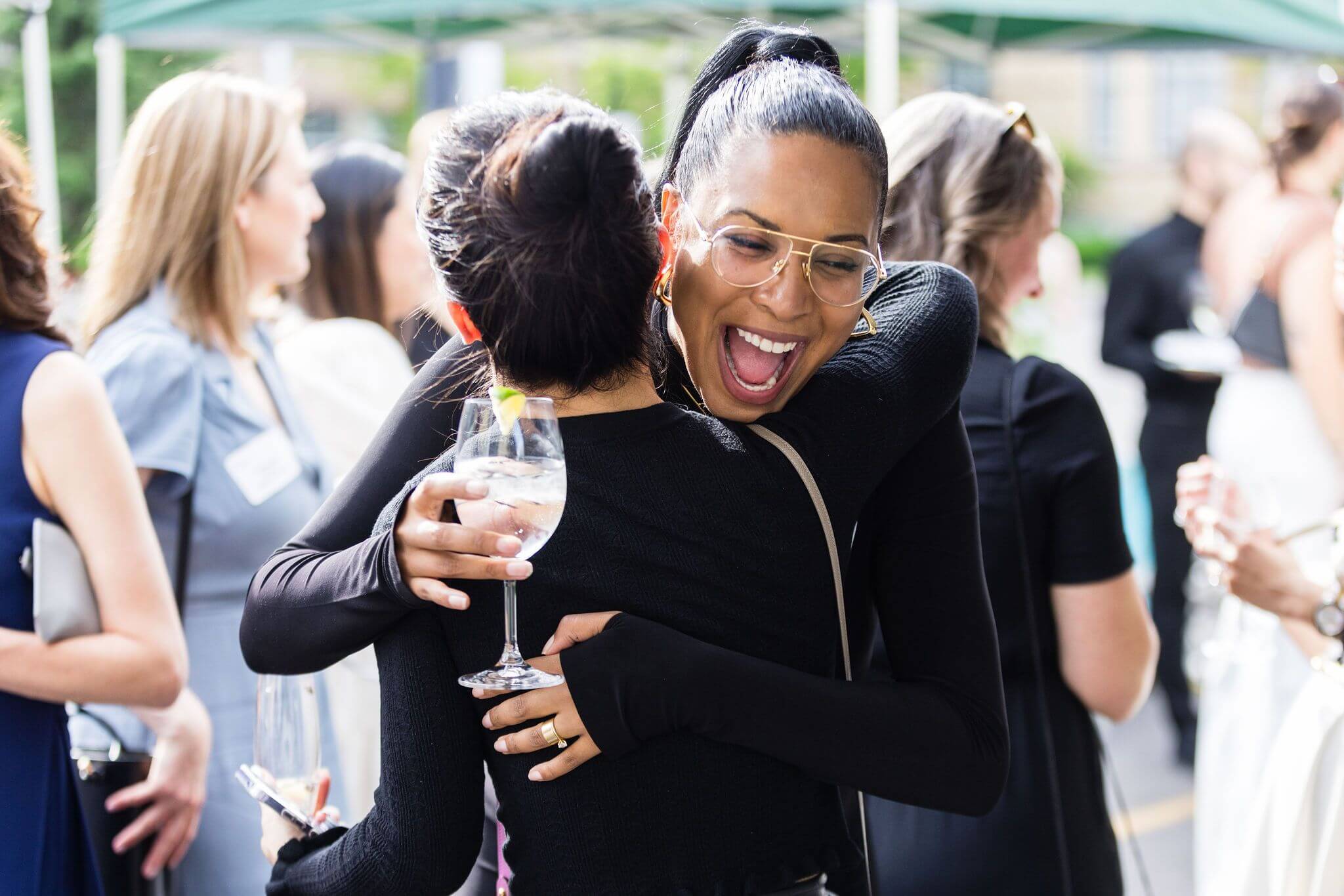 A joyful woman wearing glasses hugs another woman at an outdoor event on the Urban campus Toronto, smiling widely. The woman being hugged holds a glass of water while others mingle in the background, highlighting global education for girls.