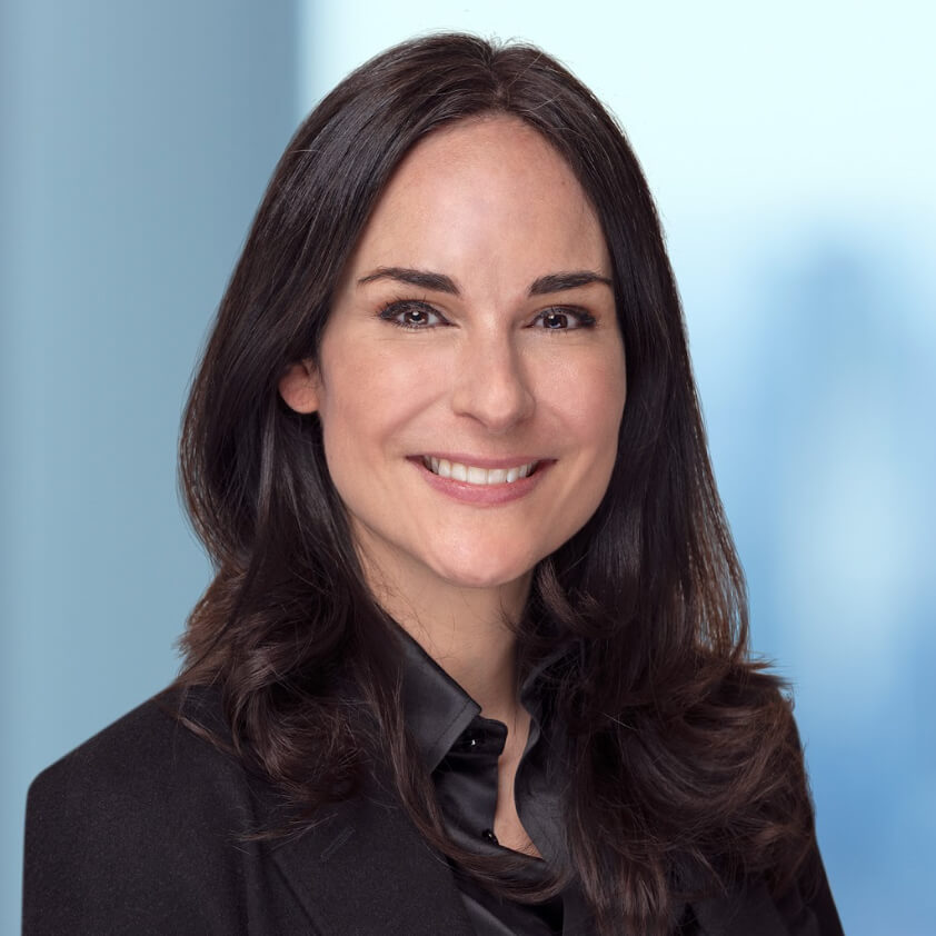 A woman with long dark hair wearing a black blazer and black shirt smiles at the camera against a light blue, blurred background.