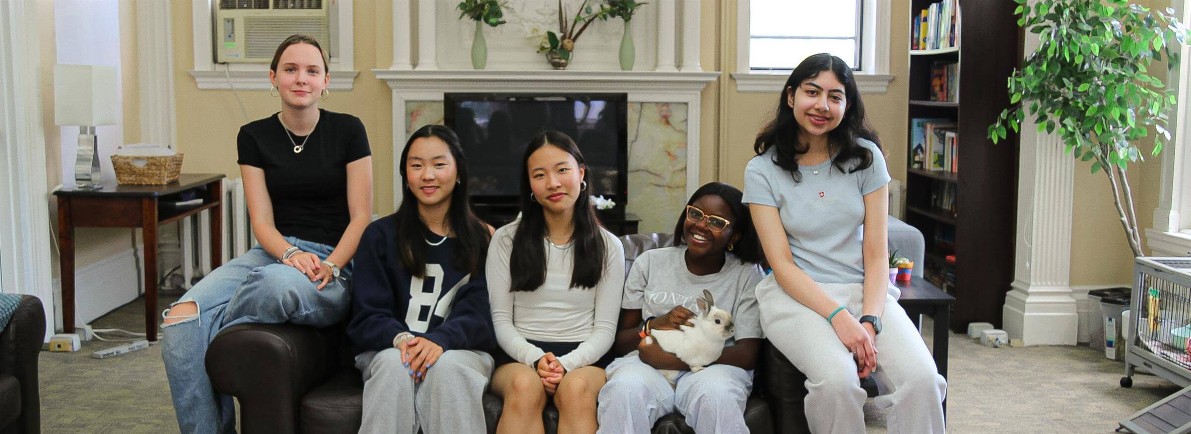 Five young people sit on a couch in a cozy room with bookshelves, a plant, and a fireplace. One person holds a white rabbit. Smiling at the camera, they embody the spirit of global education for girls found on our urban campus in Toronto.