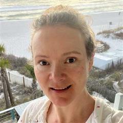 A woman with light skin and light brown hair pulled back smiles at the camera. Behind her is a beach scene with sand, greenery, fencing, and the ocean—a perfect setting for global education for girls.