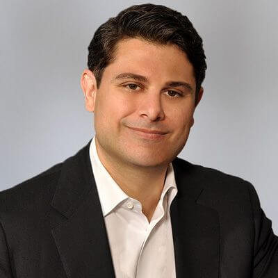 A man with short dark hair wearing a black suit jacket and white dress shirt, smiling softly against a light gray background, represents an advocate for global education for girls and supports scholarships for girls on the urban campus Toronto.