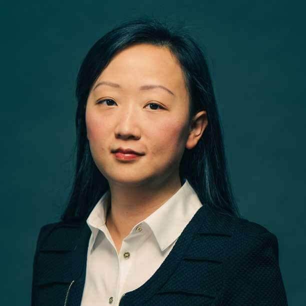 A woman with long dark hair wearing a white button-up shirt and a black blazer stands confidently before a dark green background, embodying the ambition found in the IB Diploma Programme on an urban campus in Toronto.