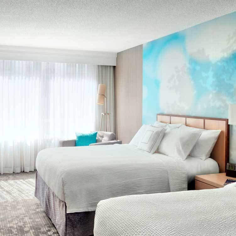 A bright hotel room features two double beds with white bedding, a blue patterned accent wall, and decor inspired by the vibrant spirit of an urban campus in Toronto. A window with sheer curtains, gray armchair, and standing lamp complete the inviting space.