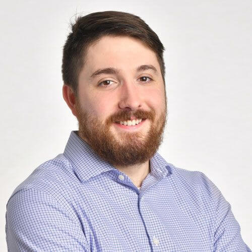 A man with brown hair and a beard, wearing a light blue checkered shirt, smiles with his arms crossed against a plain white background, proudly supporting global education for girls.