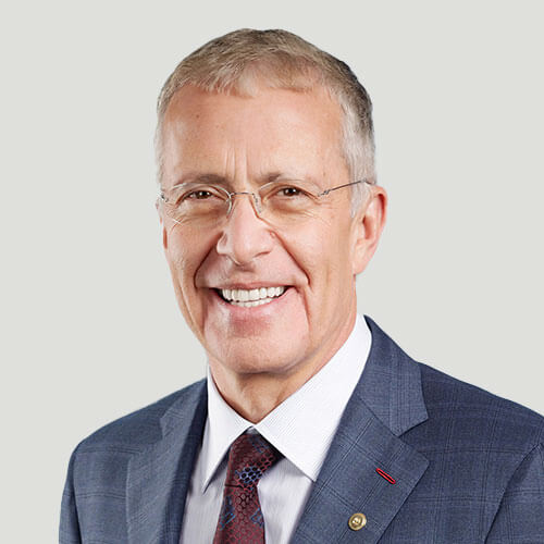 Smiling older man with short gray hair and glasses, wearing a blue checked suit, white shirt, and maroon tie, posed against a plain light background.