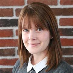 A woman with straight auburn hair and bangs is smiling slightly, wearing a white collared shirt under a gray sweater, standing in front of a brick wall—she champions global education for girls.
