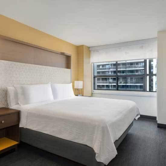 A modern hotel room with a large bed in white linens, two bedside tables with lamps, and a window overlooking tall buildings—perfect for students pursuing the IB Diploma Programme or attending global education for girls conferences. Minimal, light-colored decor adds comfort.