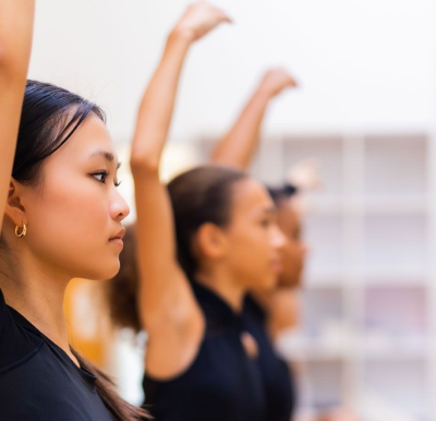 A group of young women standing in a row indoors, raising one arm, participating in what appears to be a dance or fitness class—highlighting the positive impact of global education for girls. The focus is on the woman in the foreground.