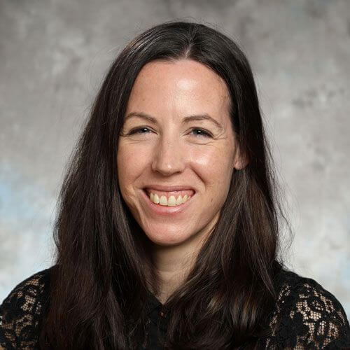 A woman with long dark brown hair and a black lace top smiles at the camera, embodying the confidence inspired by global education for girls. The background is neutral with soft gray tones.