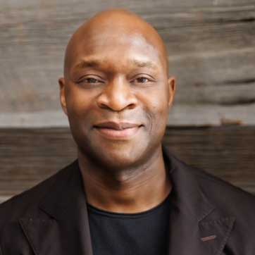 A man with dark skin and a shaved head smiles slightly while wearing a black jacket and shirt, standing in front of a wooden background.