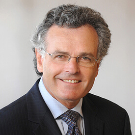 A middle-aged man with curly gray hair and glasses, smiling, wearing a dark suit, light blue shirt, and patterned tie, posed against a plain light background.