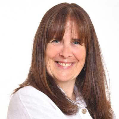 A woman with straight, long brown hair and bangs smiles at the camera. She is wearing a white collared shirt and is posed against a plain white background, reflecting the confidence inspired by global education for girls.
