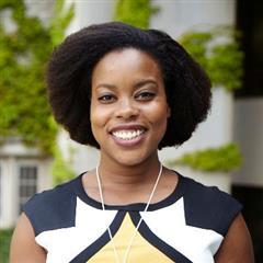 A woman with natural, shoulder-length hair smiles at the camera. She is wearing a geometric black, white, and yellow top and a necklace. The background features an urban campus Toronto building with green foliage.