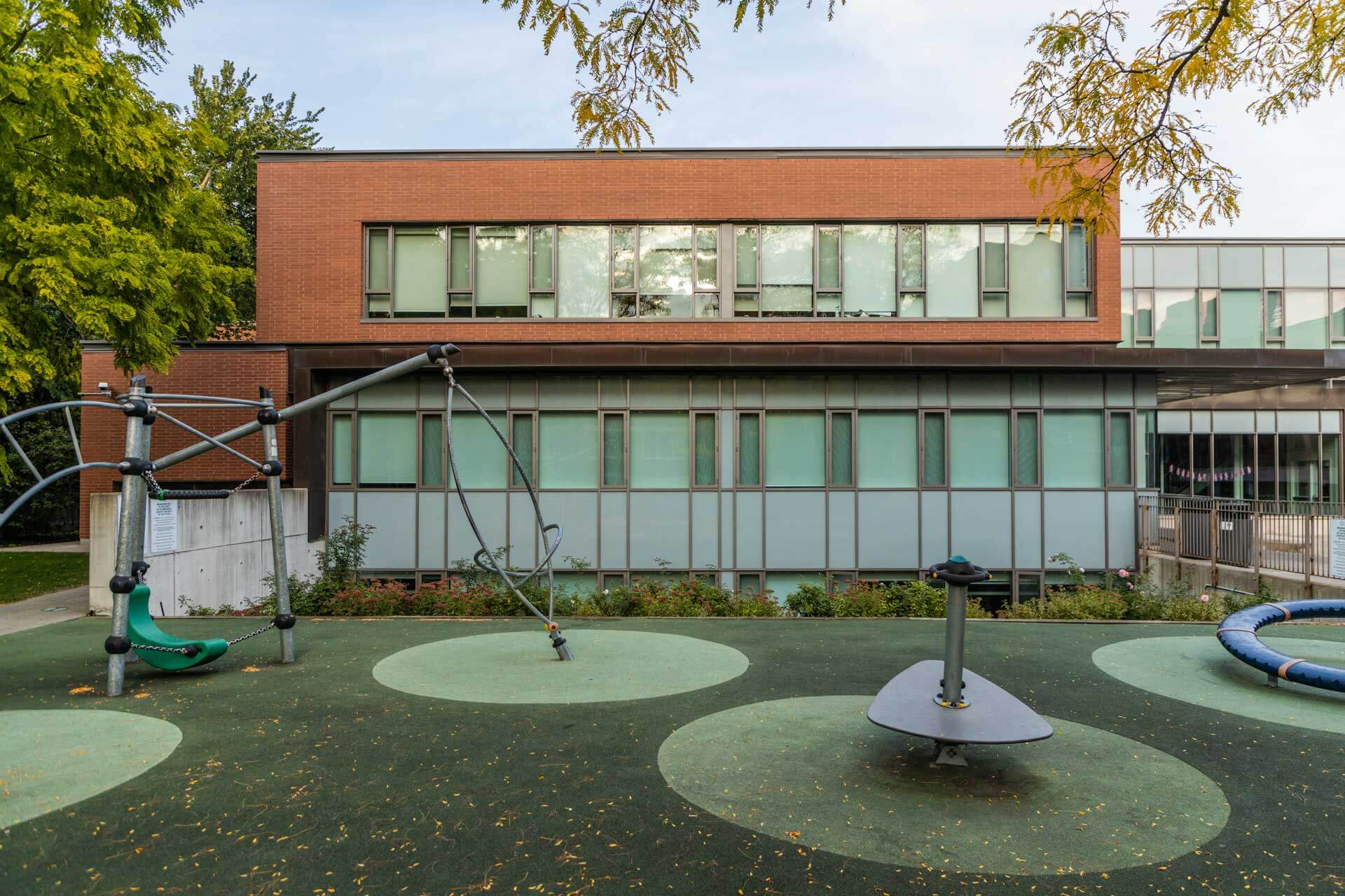 A modern playground with climbing equipment and a merry-go-round sits in front of a contemporary, two-story brick and glass building, part of an urban campus Toronto offers for global education for girls.